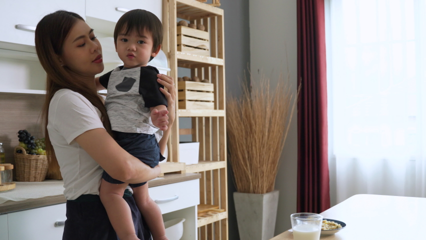 Asian young mother is holding and kissing her adorable little son affectionately in the kitchen at house.