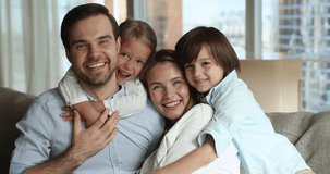 Happy attractive couple of parents piggybacking two sweet little kids, sitting on home couch, looking at camera with toothy smiles, laughing. Small children hugging mom and dad