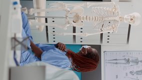 Vertical video: Old woman and nurse with face masks analyzing human skeleton bones at osteopathy consultation. Medical worker explaining orthopedic diagnosis on anatomy spinal cord at checkup visit.