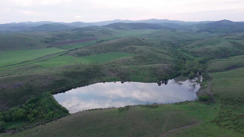 A small swampy mountain lake with clear dark cool water is located between the hills greened with fresh spring new grass, in the Balkan mountains under a cloudy blue sunny sky. UHD 4K video realtime
