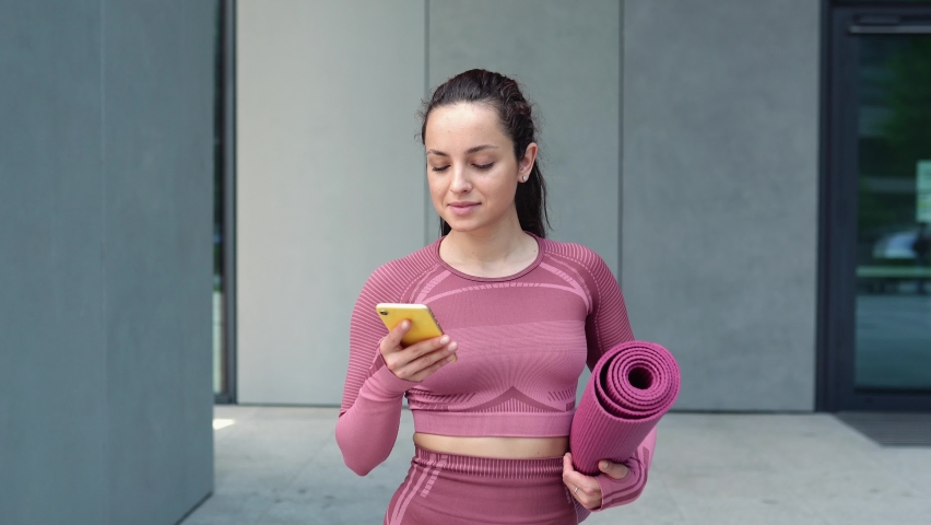Smiling fit young woman using cell phone checking fitness applications standing on urban grey wall background outdoors. Online gym free exercises, sport and health care commercial apps concept. Royalty-Free Stock Footage #1093719355