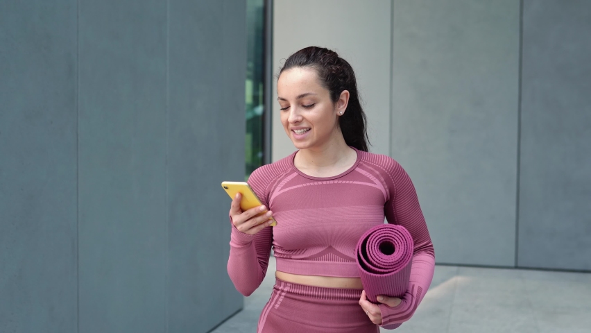 Smiling fit young woman using cell phone checking fitness applications standing on urban grey wall background outdoors. Online gym free exercises, sport and health care commercial apps concept. | Shutterstock HD Video #1093719355