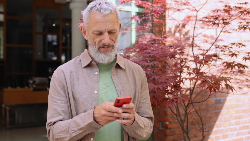 Smiling grey-haired older middle aged bearded man using moble phone outdoors. Happy old senior adult male user holding cellphone texting on smartphone, making bank payment or buying online. Royalty-Free Stock Footage #1093719367