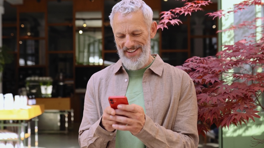 Smiling grey-haired older middle aged bearded man using moble phone outdoors. Happy old senior adult male user holding cellphone texting on smartphone, making bank payment or buying online. Royalty-Free Stock Footage #1093719367