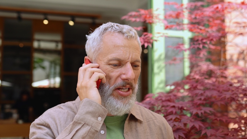 Happy european gray-haired older mid age bearded man talking on the phone outdoors. Smiling old senior adult male entrepreneur using cellphone making mobile call standing on city street outside. Royalty-Free Stock Footage #1093719375
