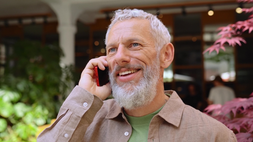 Happy european gray-haired older mid age bearded man talking on the phone outdoors. Smiling old senior adult male entrepreneur using cellphone making mobile call standing on city street outside. | Shutterstock HD Video #1093719375