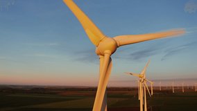 A lot of rotating wind turbines against the background of multi-colored fields in the red rays of the setting sun, a bird's eye view video from a drone. Close-up of rotating turbine blades