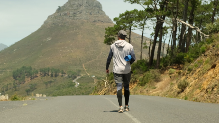 a sporty man in shorts and sports leggings walks along a road in a park near high mountains and holds a yoga mat and a jump rope in his hands. stylish sportsman man walking after jogging with yoga