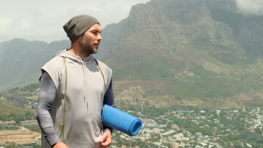 a sporty man in shorts and sports leggings walks along a road in a park near high mountains and holds a yoga mat and a jump rope in his hands. stylish sportsman man walking after jogging with yoga