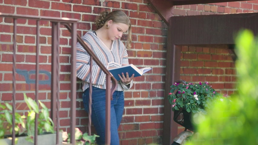 Girl reads book while leaning against a brik wall and stairs next to a flower decorated bike. Slow motion. Royalty-Free Stock Footage #1093723273