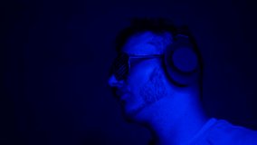 Musical background in neon blue with a man in headphones and creative club glasses. A young man listens to music with over-ear headphones