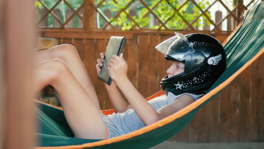 A boy lying in a hammock is playing on a tablet. A child in a motorcycle helmet enthusiastically plays racing on a tablet.