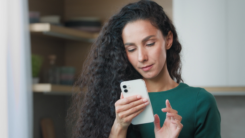 Attractive brunette woman girl with curly hair hispanic housewife look at gadget display chatting on mobile phone use smartphone for distant communication read sms from boyfriend play internet game Royalty-Free Stock Footage #1093733719