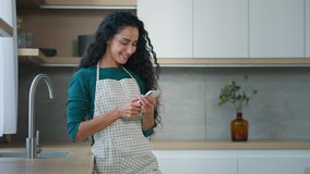 Glad smiling housewife arabian hispanic woman mother homeowner with long curly hair wears apron stand in kitchen chatting sms make cooking notes use mobile phone search internet recipe food delivery