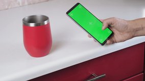 The girl is standing in the kitchen, drinking coffee and flipping through the phone screen. A woman is holding a phone with a green screen. Caucasian woman reads the news in the phone.