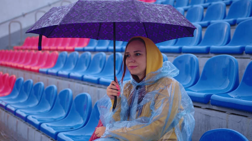 Thoughtful young female in hoodie and plastic raincoat looking away while sitting on tribune with umbrella in hand during match