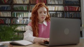 Focused Girl with red hair student using laptop search information internet course study online e learning in app typing on computer make notes write essay prepare for test exam sit at library desk