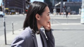 Young happy busy professional Asian business woman executive wearing suit making call on cellular holding smartphone talking on mobile phone having chat on cellphone walking on urban city street.