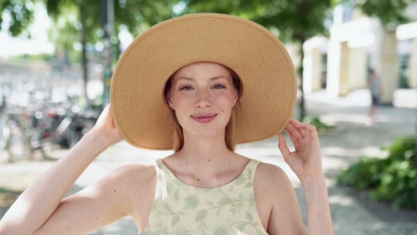 Young happy blond woman wearing hat standing on summer city street. Cheerful pretty lady fashion model looking at camera winking, dreaming of vacation, advertising accessories store outdoor.