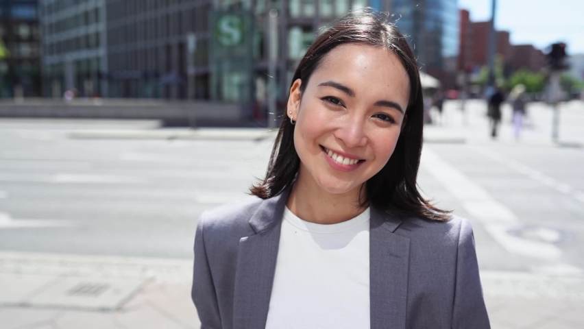 Young happy successful Asian business woman, female chinese professional executive, ethnic smiling businesswoman wearing suit standing on big city street outdoors looking at camera, close up