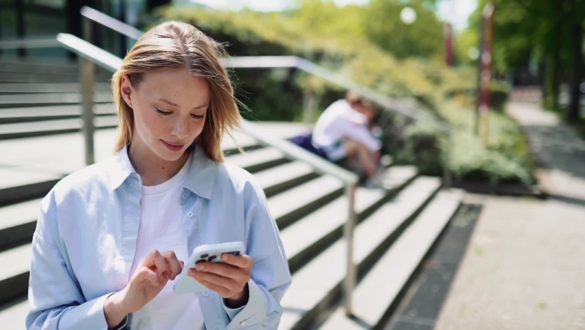 Happy pretty girl university student user holding cell phone using education tech apps looking at smartphone texting messages standing outside. Mobile online applications concept. Royalty-Free Stock Footage #1093742101