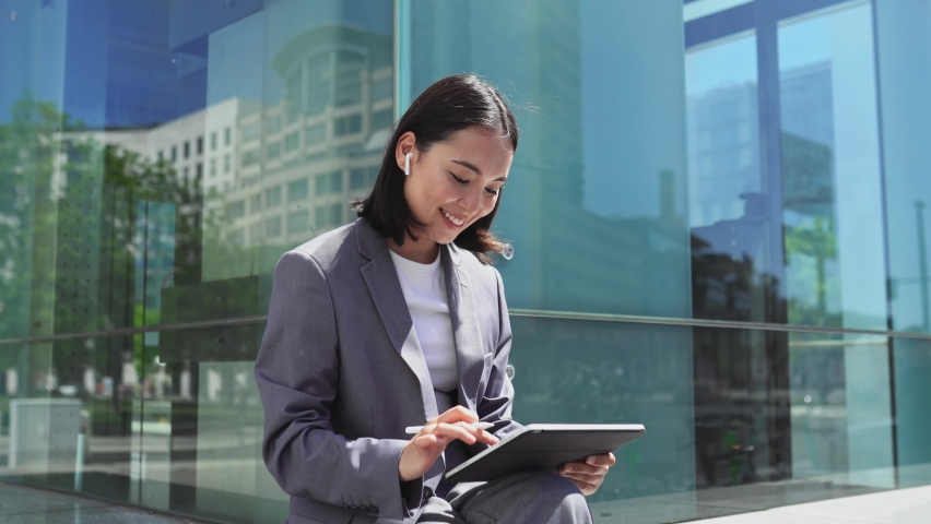 Asian elegant professional business woman wearing suit using digital tablet outside office, happy businesswoman executive holding pad corporate technology device on city street glass background.