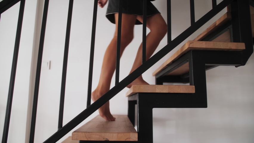 Close-up of female feet go up wooden stairs. Freelance woman walking up stairs in modern duplex apartment. Barefoot woman going up a modern wooden staircase.
