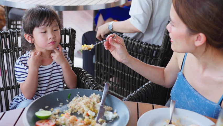 Asian beautiful mother feeding foods to young daughter in restaurant. Happy family, Attractive loving mom support and take care of little cute child kid while sit on chair for lunch at eating house. Royalty-Free Stock Footage #1093751513