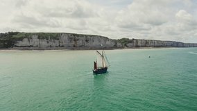 A sailing tourist ship sails along the picturesque sheer cliffs located in the outskirts of the city of Etretat. Drone bird's eye view video, France