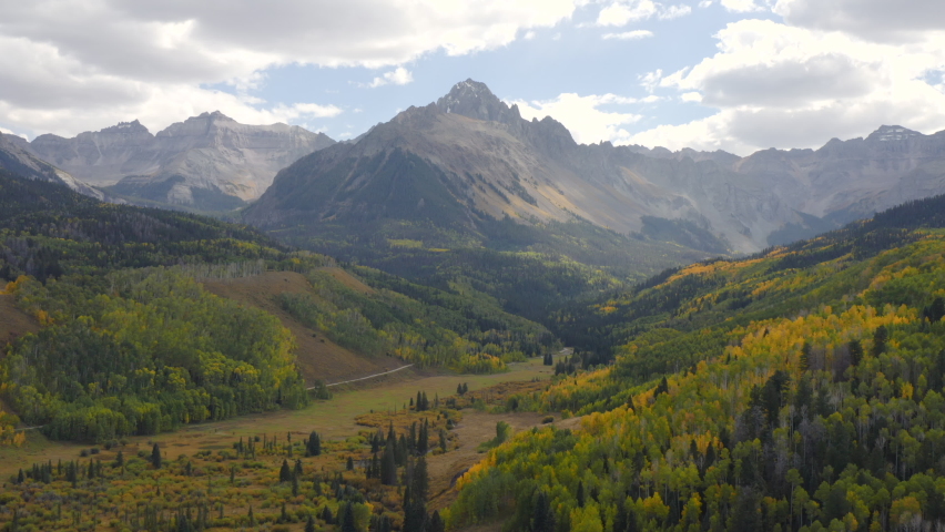 Aerial shot forward of beautiful Colorado mountains and bright yellow and orange aspen trees during autumn in the San Juan Mountains showing fall landscapes near Mt. Sneffels