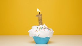 Birthday cupcake With Burning golden Candle number one on bright yellow Background. Slow motion first anniversary video banner with white confetti.