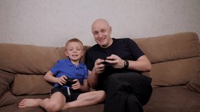 A cheerful father and his little son are sitting on the couch with joysticks in their hands playing a game. A father spends time with his son.