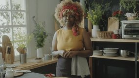 Young African American woman in apron standing in kitchen and talking on camera while filming food vlog or giving online cooking class