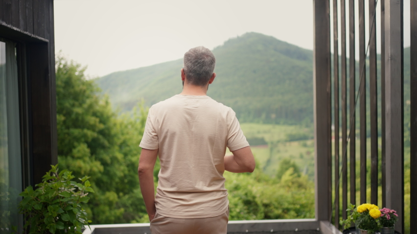 Mature man standing in tiny house terrace in front of beautiful nature with cup of coffee. Morning routine. Royalty-Free Stock Footage #1093793977