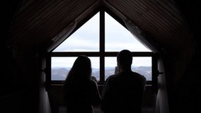 Silhouette of a young couple from behind is sitting in front of the beautiful mountains home view and drinking tea inside together.