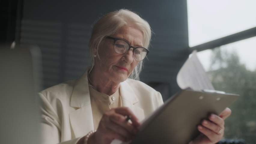 Serious older lawyer businesswoman in glasses reading signing trust partnership contract sit at table in office. Executive ceo put signature make legal bank sale financial loan investment agreement. Royalty-Free Stock Footage #1093794755