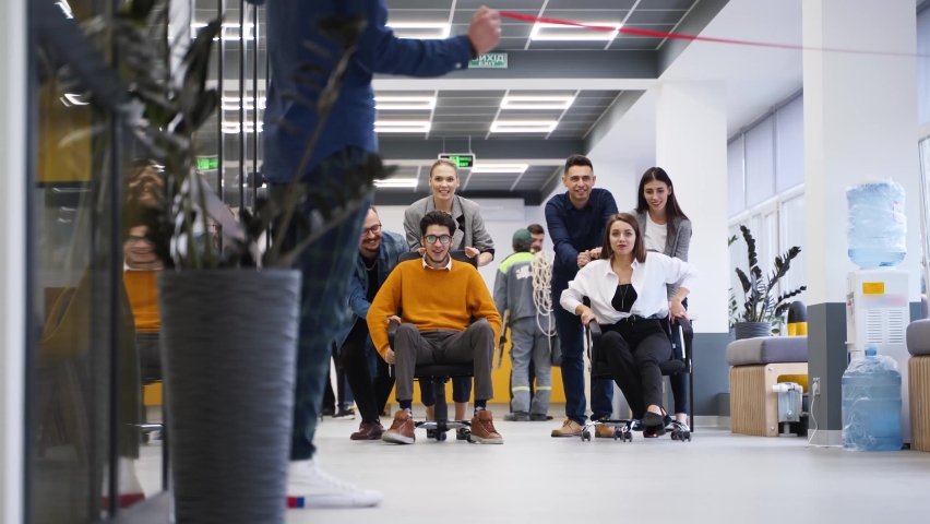 Cheerful people have fun during workday break. Two teams compete in chair on wheels racing in office hallway. Colleagues pushing coworkers on chairs. Winners rejoice. Employees film on smartphones. Royalty-Free Stock Footage #1093810187