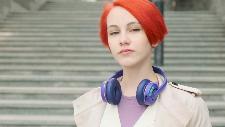 Close-up portrait of Caucasian happy beautiful girl with red hair in raincoat, standing on street looking at camera and smiling outdoors. joyful girl puts on headphones in good mood in city