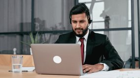 Successful confident indian or arabian businessman, company ceo, with headset, look at laptop screen, waves hand, talking with business client or partner online, explain business strategy and plan