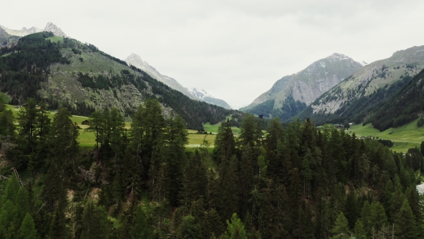 The drone rises from behind the trees, revealing beautiful panoramic view of high mountain valley with a village in the lowland. The peaks of the rocky Alpine mountains are covered with snow