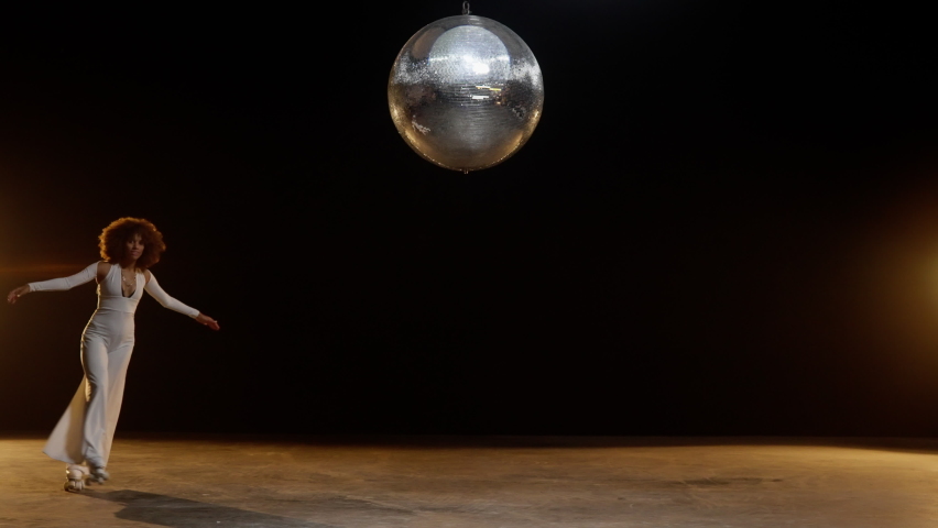 Charming African American Woman with Fluffy Afro Style Hairs in White Jumpsuit Wearing Roller Skates Rides Around on the Stage Under Shiny Disco Ball. Cinematic Slow Motion Shot