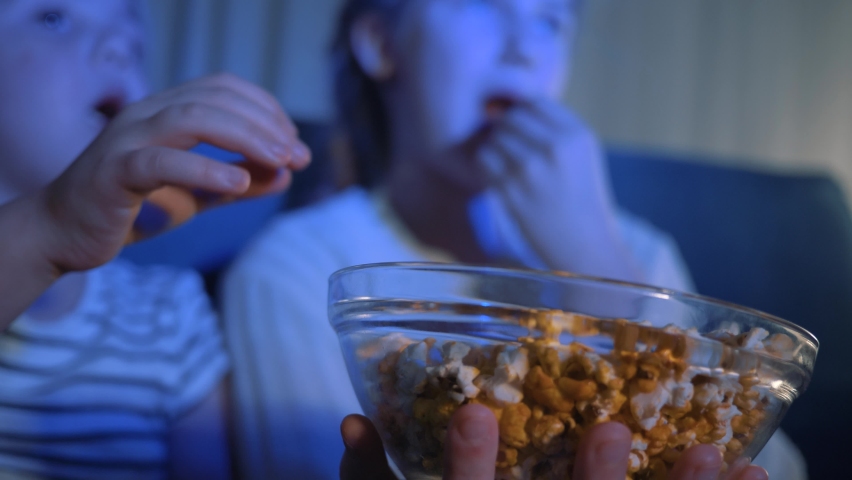 Plate is submissive in hands. Sister girls are watching TV. eating popcorn sitting on couch. Children take popcorn from plate in front of TV eat. Children watch their favorite TV, eat salted popcorn Royalty-Free Stock Footage #1093840351