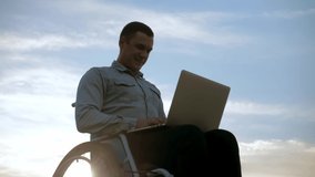 Young man in a wheelchair works at his laptop sitting against a beautiful sky.Disabled guy works at his laptop on the beach. Works remotely.