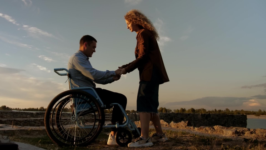 Young man in a wheelchair trying to get up. A girl helps an invalid get up. A  man stands up against a beautiful sunset Royalty-Free Stock Footage #1093841431