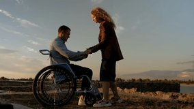 Young man in a wheelchair trying to get up. A girl helps an invalid get up. A  man stands up against a beautiful sunset