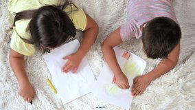 Siblings playing together at home. Top view of little boy and girl lying on the carpet and drawing on white sheets of paper with colorful crayons. High quality 4k footage