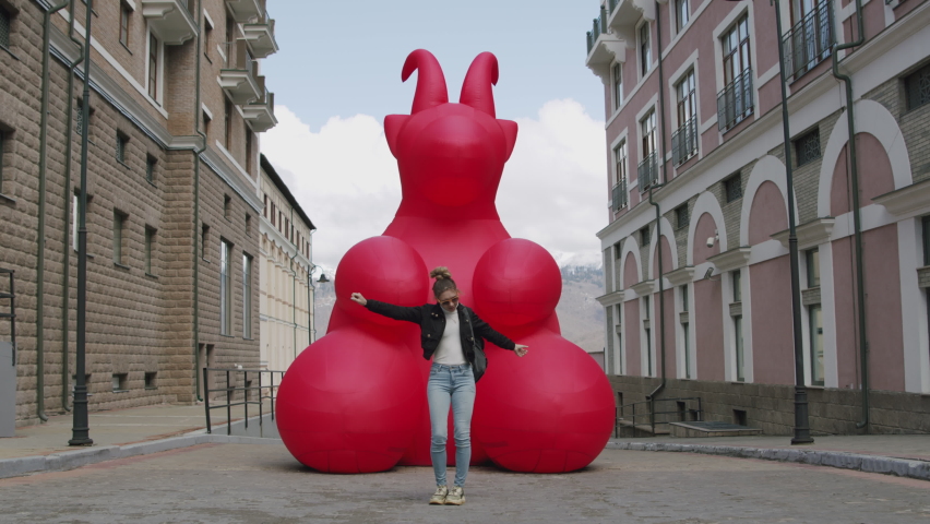 A cheerful fashionable multi-racial girl in yellow sunglasses is dancing funny and fun on the street in front of a huge red inflated goat toy. A village in the Alps