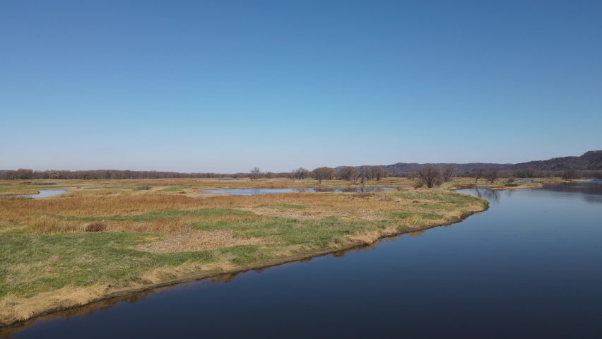 View over water ways in Wisconsin on a bright, clear autumn day. Long grasses drying on the intermittent island. Sun casting reflections off the calm water. Bare trees in clumps on the islands, 