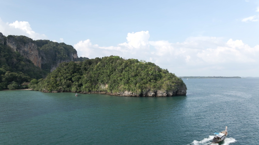 Small hopping tour boat traveling along the Railay peninsular isle coastline in Krabi province of Thailand - Aerial tracking shot 