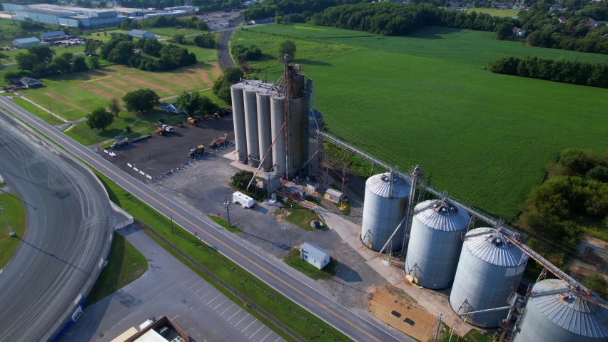 An aerial view of silos on a farm on a sunny day. The camera tilted down, truck right and pan left orbiting the silos. Then a horse track is revealed in the background, along with warehouses.